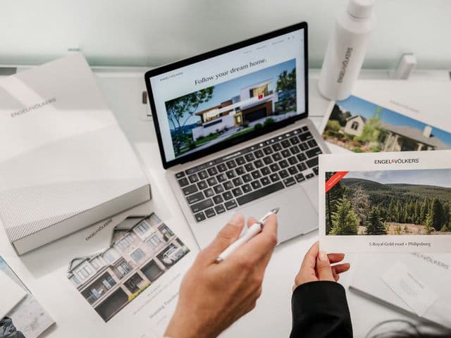 Overhead view of a desk with a laptop displaying luxury property listing, hands holding property photos, printed marketing materials, and property brochures in a clean white workspace setting