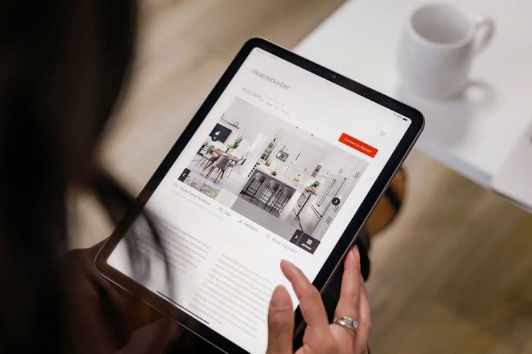 Close-up view of hands holding a tablet displaying a real estate website with property listings and interior photos, with a white coffee cup in the background