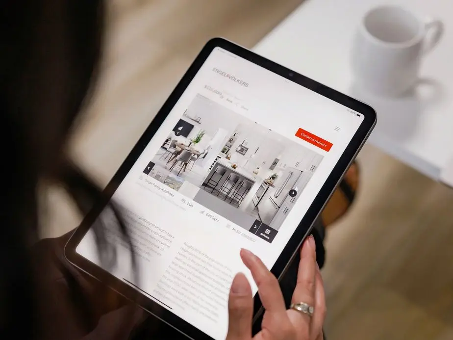 Close-up view of hands holding a tablet displaying a real estate website with property listings and interior photos, with a white coffee cup in the background