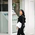 A female real estate agent in a black business suit carrying documents while walking past an office window display featuring property listings. The image captures a professional moment in a modern real estate office setting.