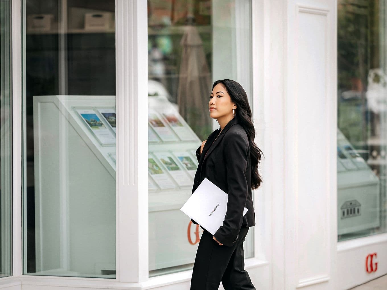A female real estate agent in a black business suit carrying documents while walking past an office window display featuring property listings. The image captures a professional moment in a modern real estate office setting.