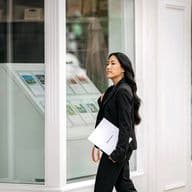 Real estate agent in black suit walks past a white building with property listings in the window. She carries a white Engel & Volkers folder.