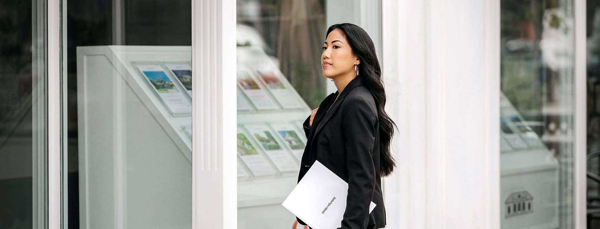 Real estate agent in black suit walks past a white building with property listings in the window. She carries a white Engel & Volkers folder.