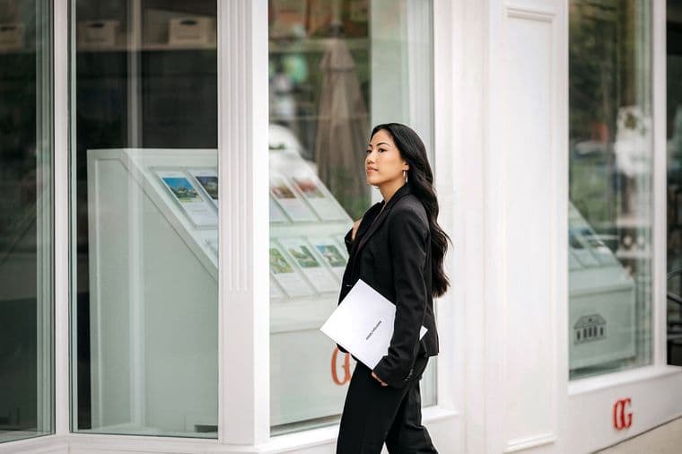 Une agente immobilière en costume noir passe devant un bâtiment blanc dont la vitrine affiche des annonces immobilières. Elle porte un dossier blanc Engel & Volkers.