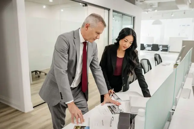 Two real estate professionals collaborating at a desk in a bright modern office space. A man in a gray suit with a red tie and a woman in a black blazer are leaning over documents and materials spread on a white desk, engaged in discussion.