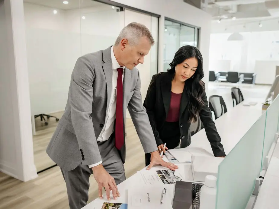 Two real estate professionals collaborating at a desk in a bright modern office space. A man in a gray suit with a red tie and a woman in a black blazer are leaning over documents and materials spread on a white desk, engaged in discussion.