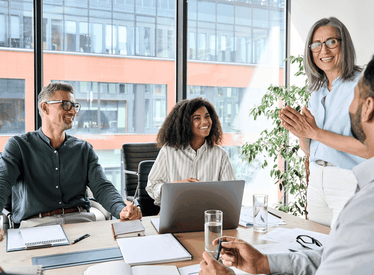 A diverse group of professionals are gathered around a conference table in a modern office, smiling and engaged in a meeting.