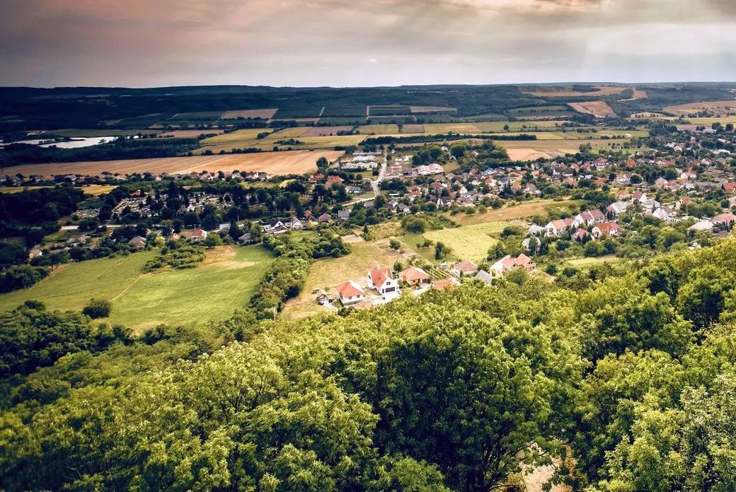 Aerial view of a rural landscape around Pest, lush green fields, and distant hills under a cloudy sky with sun rays.