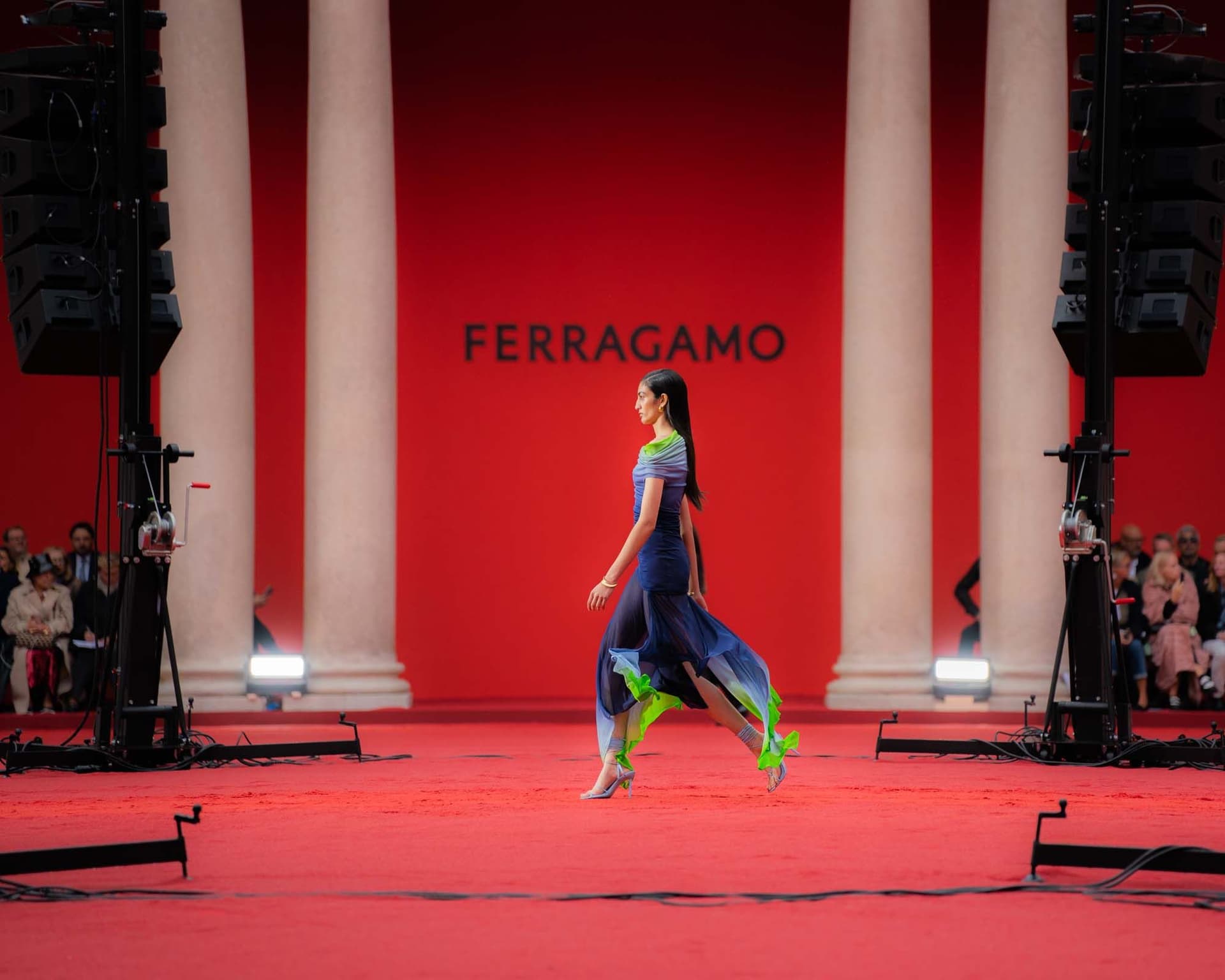 A model in a blue-green, flowing dress walks down a red catwalk with the word ‘FERRAGAMO’ in the background, flanked by large columns.