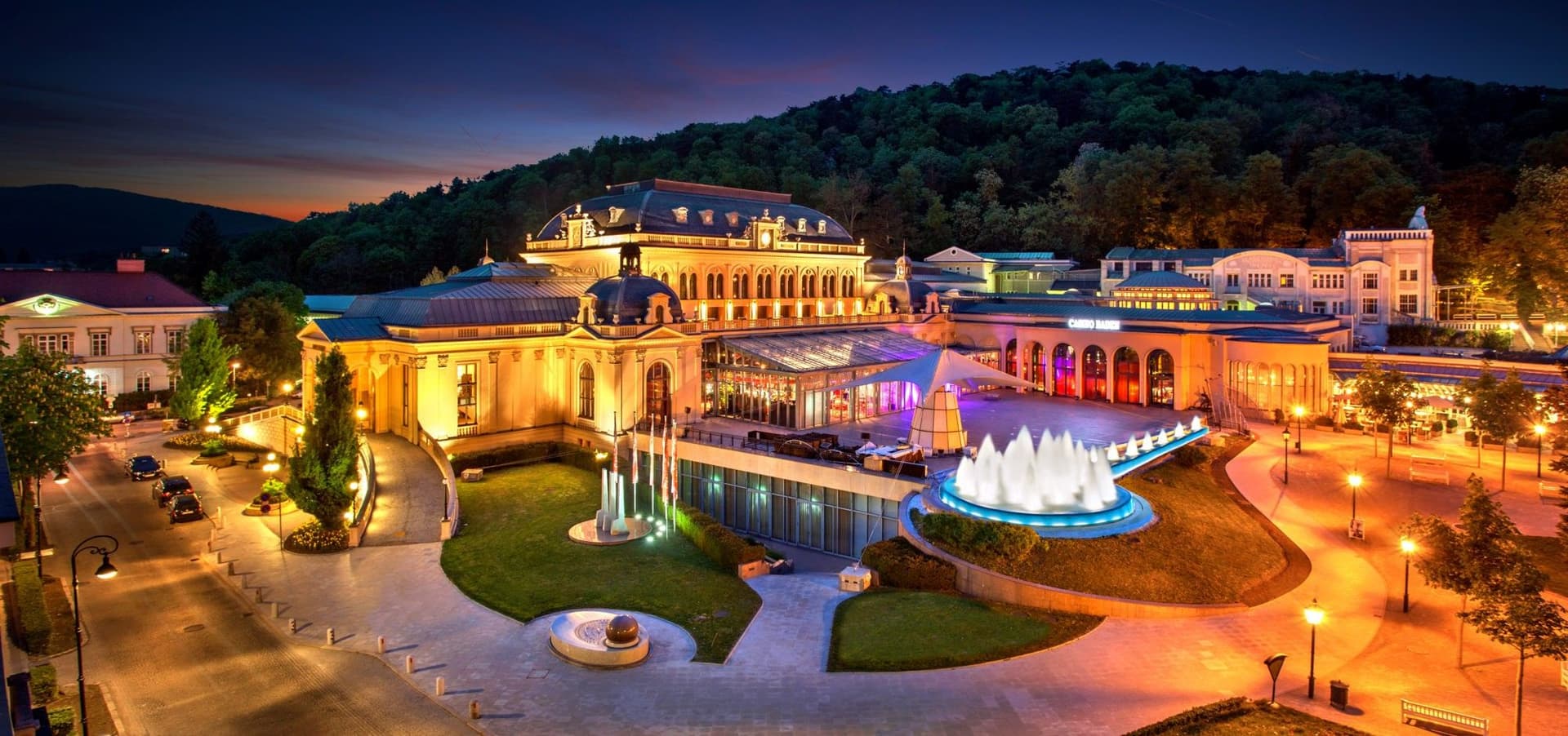 Panoramaaufnahme des Casino Baden in der Dämmerung, mit beleuchteter historischer Fassade, Springbrunnen und Parkanlage in warmen Gold- und Blautönen