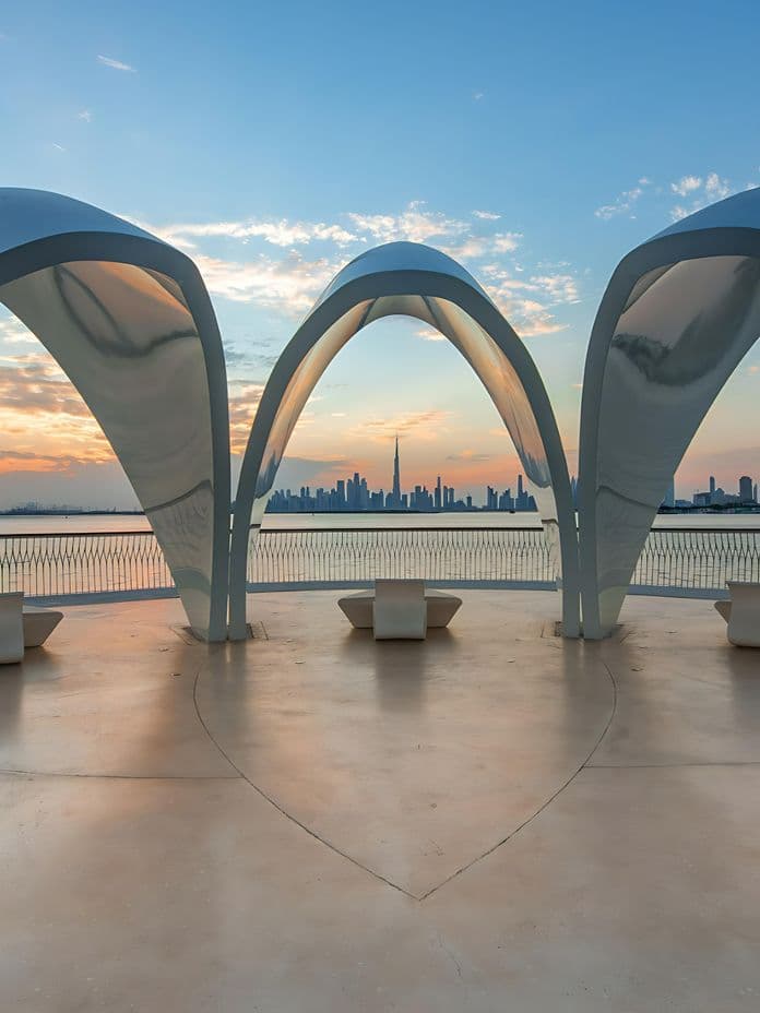 Modern architectural structure with curved arches overlooking Dubai skyline at sunset, featuring Burj Khalifa in the distance.