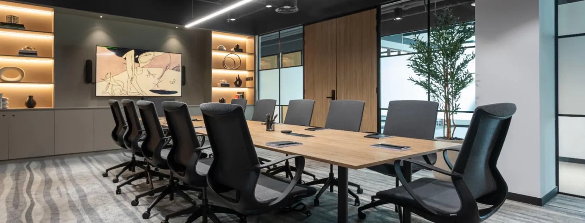 Modern conference room with a long wooden table, black swivel chairs, shelves with decor, and a large artwork on the wall.