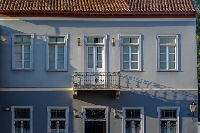 Zweigeschossiges graues Gebäude mit einem roten Ziegeldach. Eine schwarze Tür befindet sich mittig im Erdgeschoss. Ein Balkon befindet sich im ersten Stock.