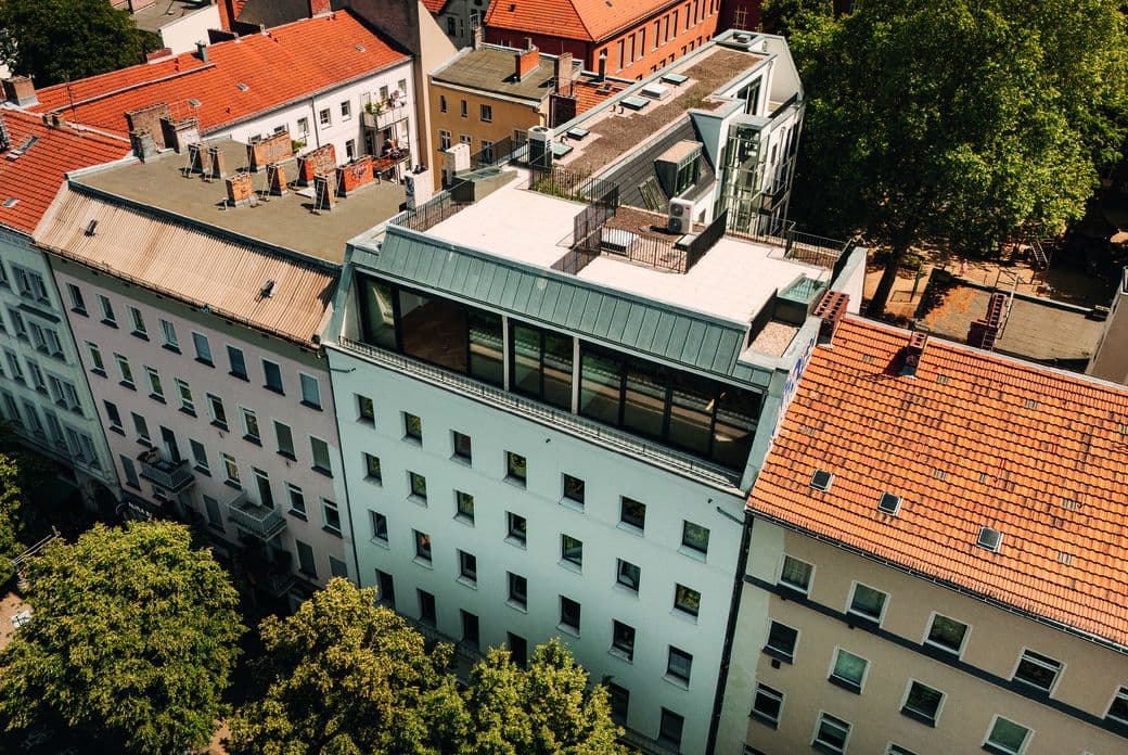 Aerial view of a city block: orange-tiled roofs, white and beige buildings, a rooftop terrace with a glass extension, and dense green trees.