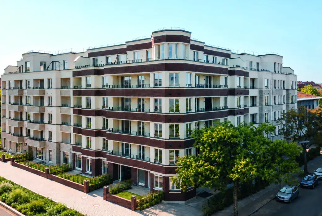 Modern white and brick apartment building with numerous balconies and glass railings, set among trees with a street and parked cars.