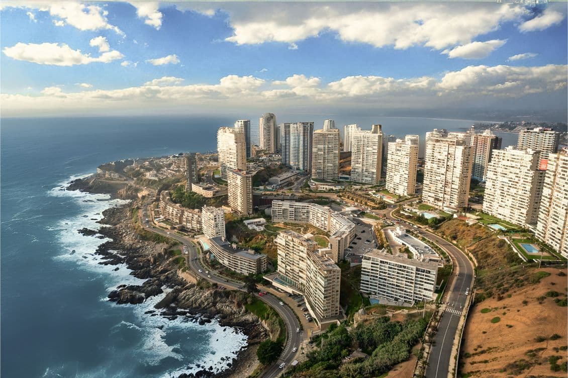 Vista aérea de una ciudad costera con edificios altos, carreteras sinuosas y una costa rocosa junto al océano bajo un cielo parcialmente nublado.