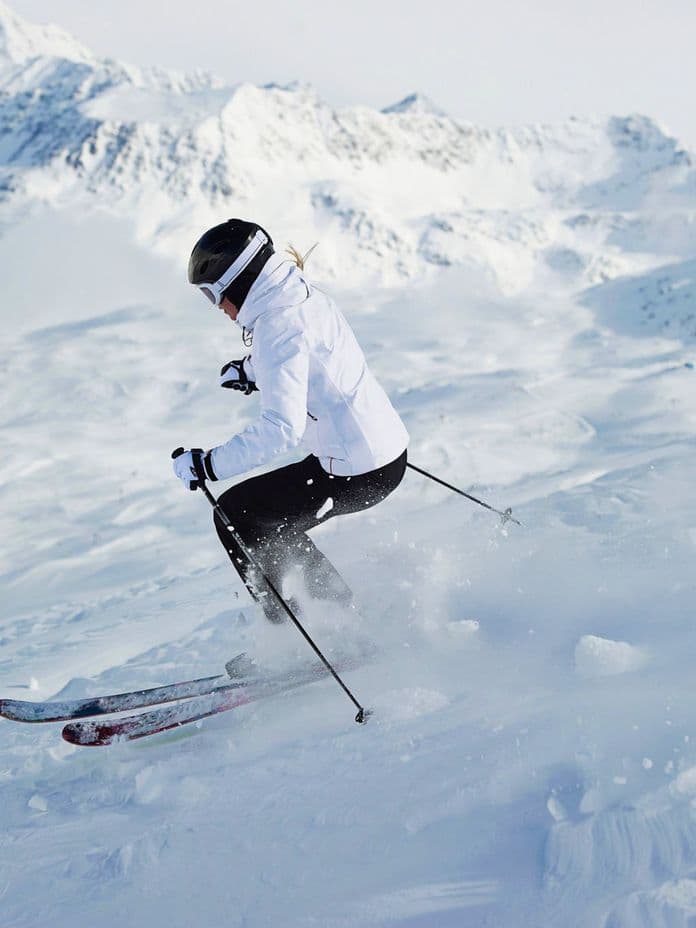 A skier in a white jacket descends a snowy slope, kicking up snow, with snow-covered mountains in the background.
