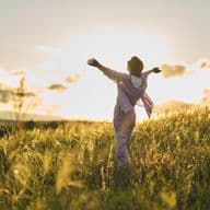 A woman stands with outstretched arms in a sun-drenched meadow, soaking in the beauty of nature in the evening light. A symbolic image of quality of life in Tirol & Zell am See.
