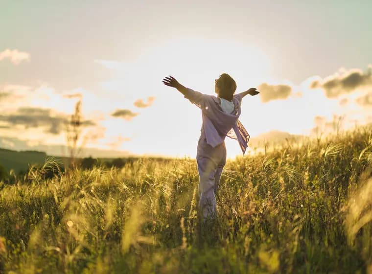 A woman stands with outstretched arms in a sun-drenched meadow, soaking in the beauty of nature in the evening light. A symbolic image of quality of life in Tirol & Zell am See.