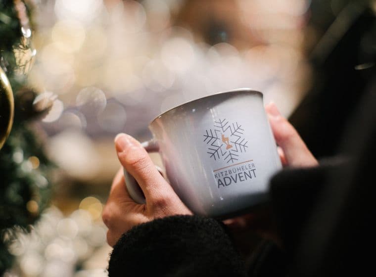 Person hält eine Tasse mit Kitzbüheler Advent Logo, im Hintergrund weihnachtliche Beleuchtung.