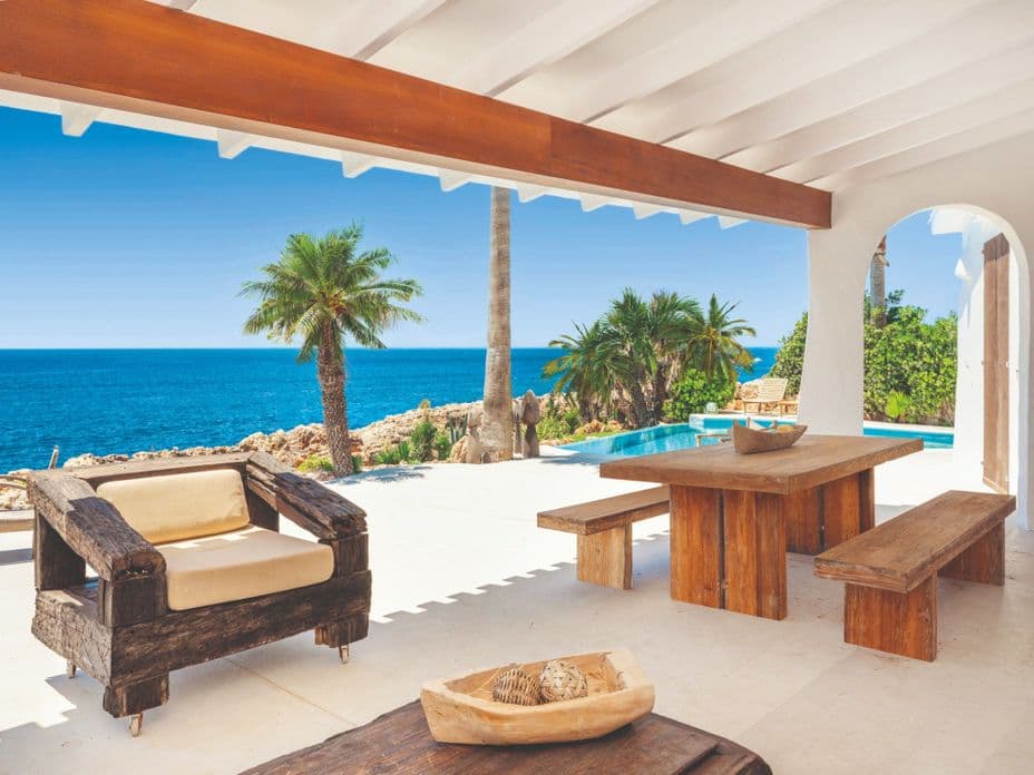 Terrassa coberta d’un allotjament de vacances a Menorca amb mobiliari rústic de fusta, palmeres, piscina privada i vistes panoràmiques al mar Mediterrani. 2/2