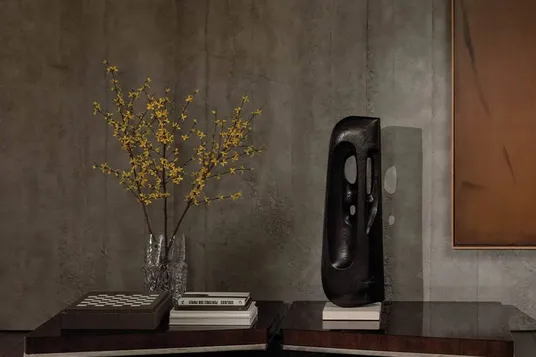 Contemporary room with an exposed concrete wall; glass vase with yellow flowering branches on a dark shelf, stacked books, a black leather chair, and a fluffy white rug.