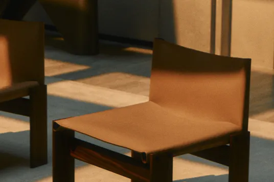 Dim room with wooden chairs on a blue carpet; a small table holds a rock, books, and a dark sculpture lit by warm light.