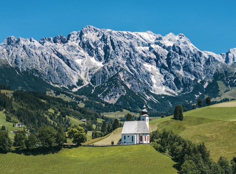 View of the Hochkönig mountain with a small church on a green meadow, surrounded by alpine landscape.