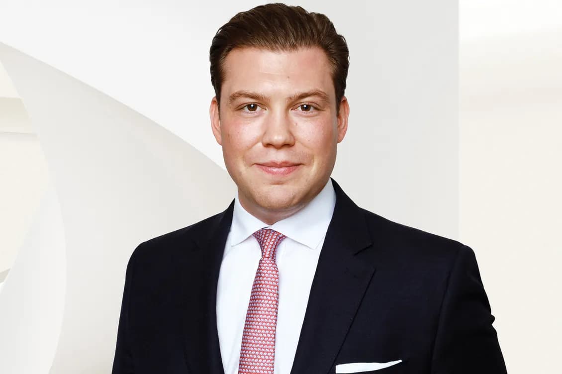 Head-and-shoulders portrait of a smiling man in a dark suit, white shirt, red patterned tie, and white pocket square, against a pale geometric background.
