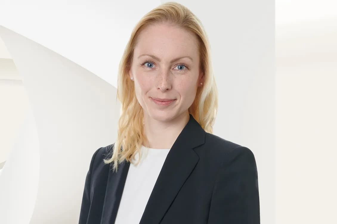Portrait of a blue-eyed blonde woman in a navy blazer and white top, against a pale geometric background.