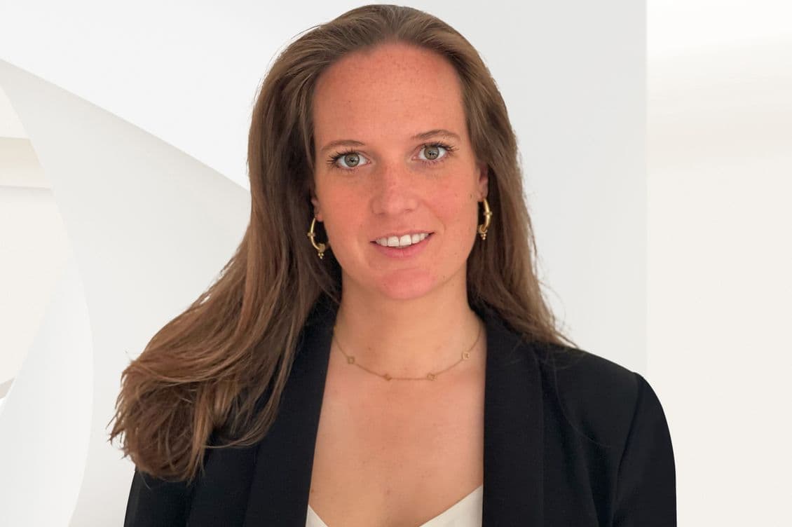 A smiling woman with long brown hair, wearing a black blazer and gold jewelry, poses against a white background.