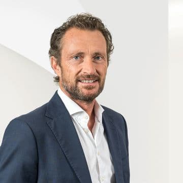 Dr. Andreas Hillig, der ein weißes Hemd und einen dunklen Blazer trägt, vor einem hellen, abstrakten Hintergrund.