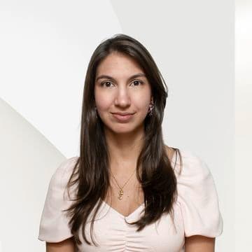 Headshot of a smiling woman with long brown hair, wearing a light pink blouse and gold necklace, against a white background.
