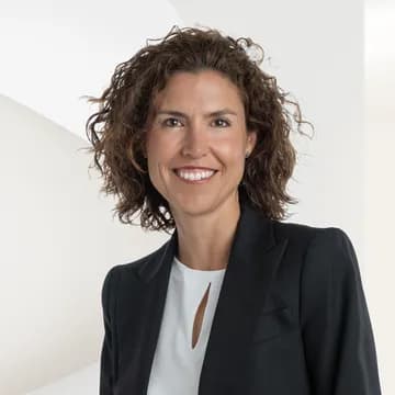 Smiling woman with short curly brown hair in a black blazer and white blouse, against a pale geometric backdrop.