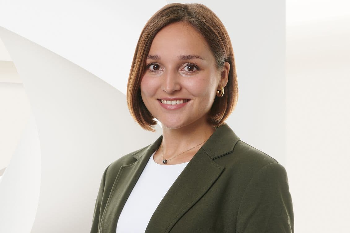 Smiling woman with short brown hair in a green blazer and white top, standing against a modern, white abstract background.