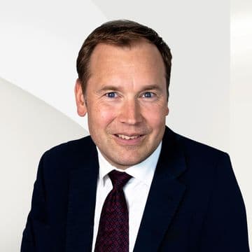 Hubertus Crasemann in Anzug und Krawatte vor einem neutralen, geometrischen Hintergrund.