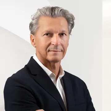 Close-up portrait of a middle-aged man with silver hair in a dark suit and white shirt, posed against a light geometric background.