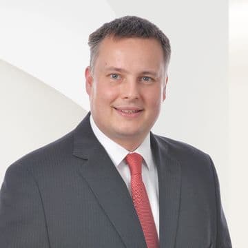 Smiling man in a dark suit and red tie with a neutral white background.