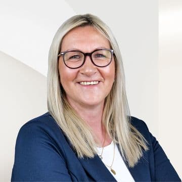 Petra Lüders mit blonden Haaren und Brille, die einen dunkelblauen Blazer über einem weißen Oberteil trägt und vor einem hellen Hintergrund steht.