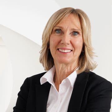 Smiling woman with blonde hair in a black blazer and white shirt, standing against a modern white abstract background.