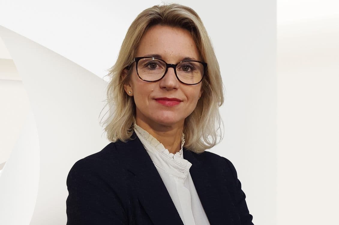 Headshot of a blonde woman with glasses, wearing a black blazer and white blouse, against a white background.