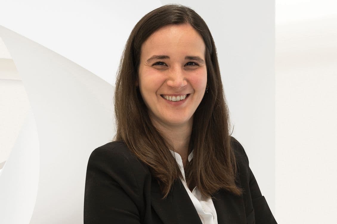 Una dona somrient amb el cabell llarg i castany, que porta una americana negra i una camisa blanca, està dreta sobre un fons blanc.