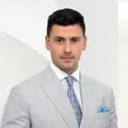 Luca Cappalonga in a light gray suit with a blue tie and pocket square, standing against a white background with geometric shapes.