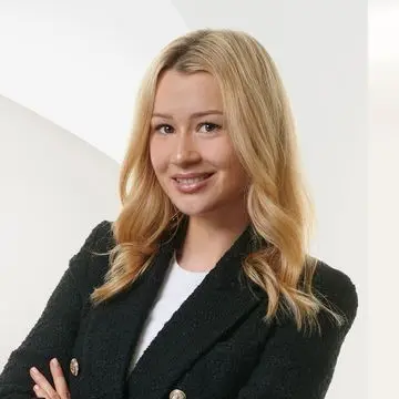 Smiling woman with long blonde hair wearing a black blazer and white shirt, standing against a white geometric background.