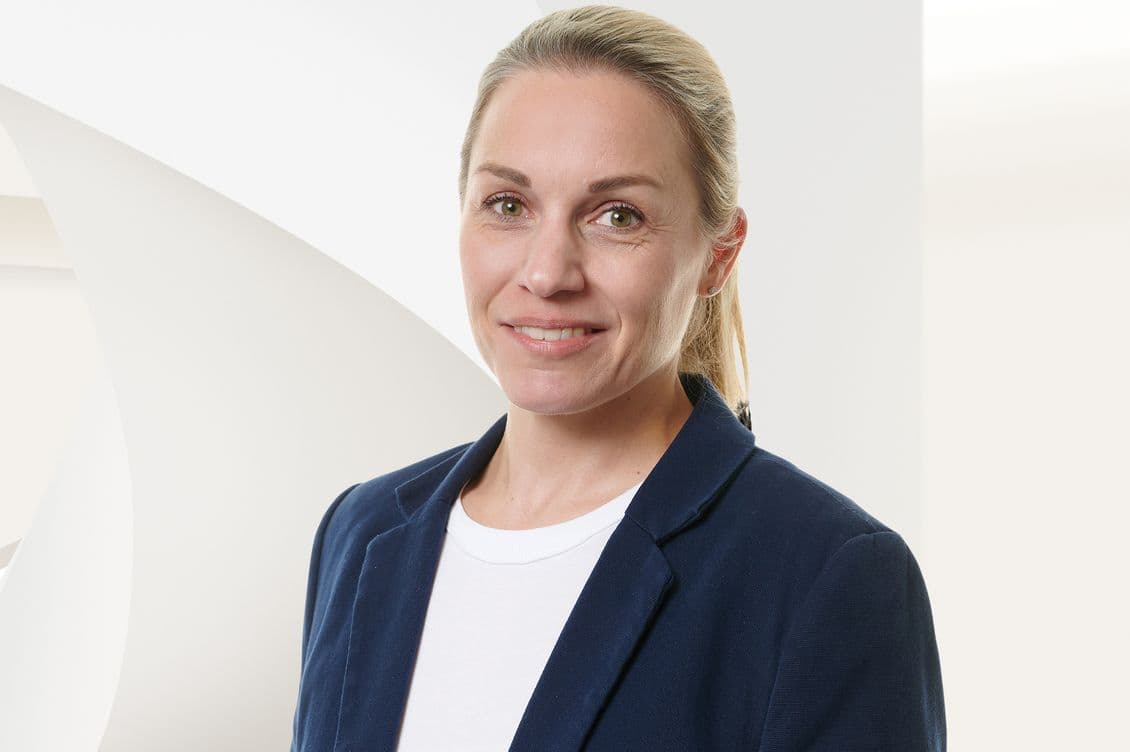 Lächelnde Frau mit blonden Haaren im Pferdeschwanz, in einem marineblauen Blazer und einem weißen Hemd, stehend vor einem hellen, abstrakten Hintergrund.