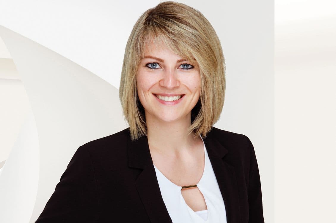 Lächelnde Person mit kurzen blonden Haaren, die einen schwarzen Blazer und eine weiße Bluse trägt, stehend vor einem weißen Hintergrund.