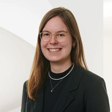 Smiling person with long hair and glasses, wearing a black blazer and necklace, standing against a white background with curved patterns.