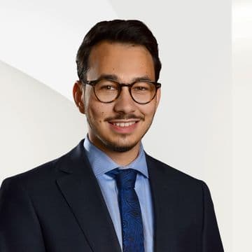 Benjamin Polewski im Anzug und mit blauer Krawatte vor einem hellen, abstrakten Hintergrund.