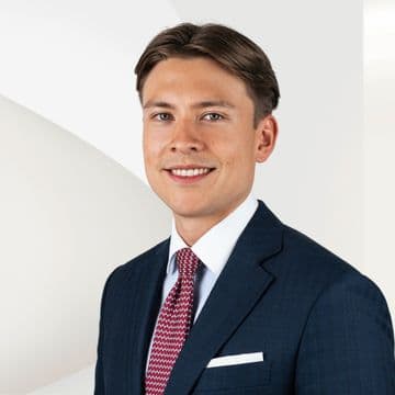 Leonardo Castillo in einem dunkelblauen Anzug und einer rot gemusterten Krawatte lächelt vor einem hellen, abstrakten Hintergrund.