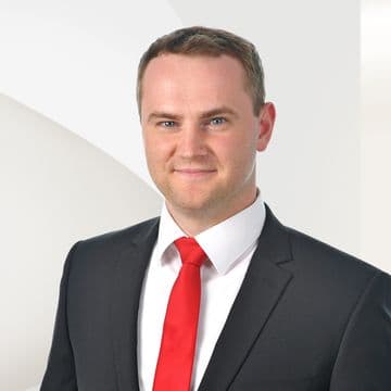 Man in a suit with a red tie, smiling against a white geometric background.